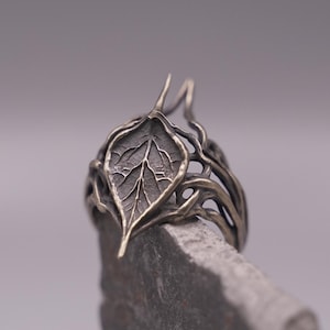 Può includere: Un anello color argento scuro caratterizzato da un motivo a foglia centrale con venature dettagliate e una superficie testurizzata. La fascia è composta da fili metallici intrecciati a forma di viticcio che creano un design traforato.