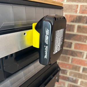 May include: A black Makita 18V 2.0Ah battery pack is mounted on a yellow holder. The battery is attached to a metal rail. The holder is mounted on a black and clear storage unit. The background is a brick wall.