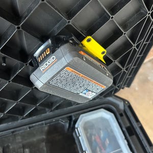 May include: A black plastic tool box with a Ridgid Lithium 1.5 battery attached to the lid. The battery is grey and black with a yellow clip. The tool box is open, revealing the interior.