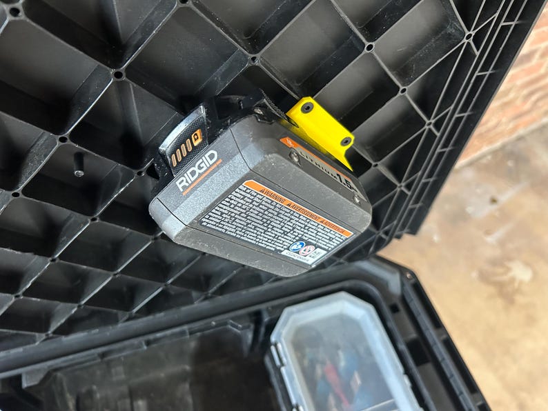 May include: A black plastic tool box with a Ridgid Lithium 1.5 battery attached to the lid. The battery is grey and black with a yellow clip. The tool box is open, revealing the interior.