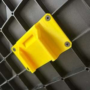 May include: Bright yellow plastic bracket with a rectangular shape, mounted on a dark gray, grid-patterned surface. The bracket is secured with three small screws. The design features a recessed area and angled sides.