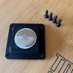 May include: A black square holder with an Apple AirTag and four black screws on a wooden surface. The AirTag is silver with the Apple logo. The holder has a textured design.
