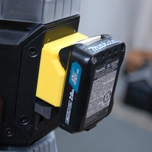 May include: Close-up of a black and yellow power tool with a black battery pack attached. The battery pack has the brand name "Makita" and the text "Li-ion 2.0Ah 12V". The tool has an orange button.
