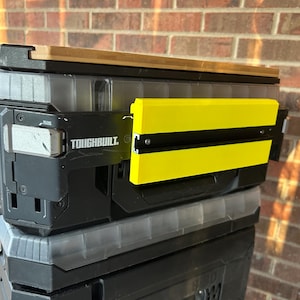 May include: A black and gray ToughBuilt toolbox with a yellow and black side panel. The toolbox has a wooden top and clear plastic compartments. The ToughBuilt logo is visible on the side.