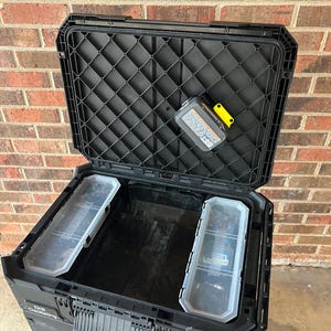May include: Open black toolbox with a textured lid and two clear plastic storage trays. A battery pack with a yellow accent is inside the lid. The toolbox is open, revealing the interior.