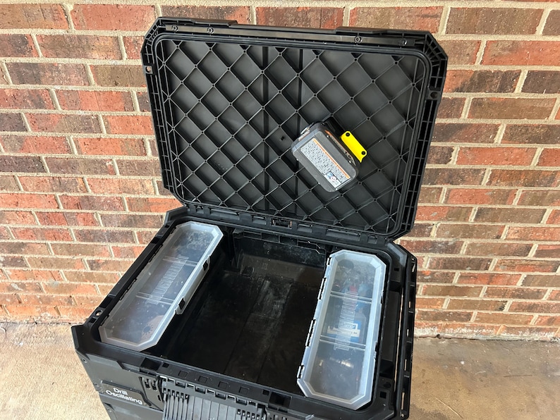 May include: Open black toolbox with a textured lid and two clear plastic storage trays. A battery pack with a yellow accent is inside the lid. The toolbox is open, revealing the interior.