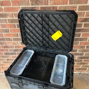 May include: Black open tool box with a diamond pattern on the lid. The tool box has two clear plastic storage containers inside. A yellow plastic piece is attached to the inside of the lid. The tool box is in front of a brick wall.