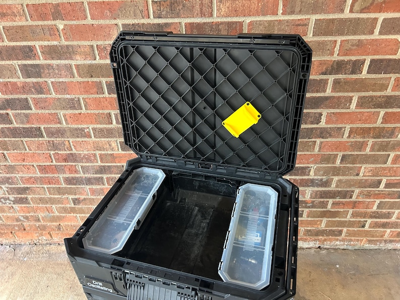 May include: Black open tool box with a diamond pattern on the lid. The tool box has two clear plastic storage containers inside. A yellow plastic piece is attached to the inside of the lid. The tool box is in front of a brick wall.
