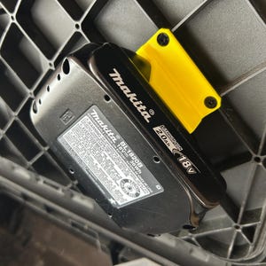 May include: Black Makita BL1820B 18V 2.0Ah lithium-ion battery with a yellow plastic holder. The battery is labeled with warnings and specifications. The battery is placed on a black plastic grid.