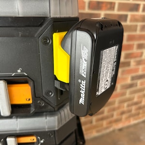 May include: A black Makita 18V 2.0Ah lithium-ion battery attached to a black and yellow tool. The battery is mounted on a yellow bracket. The background is a brick wall.