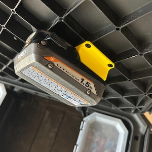 May include: A Ridgid 18V Lithium 1.5Ah battery pack with a yellow holder attached to a black plastic storage container. The battery is grey and black with white text.