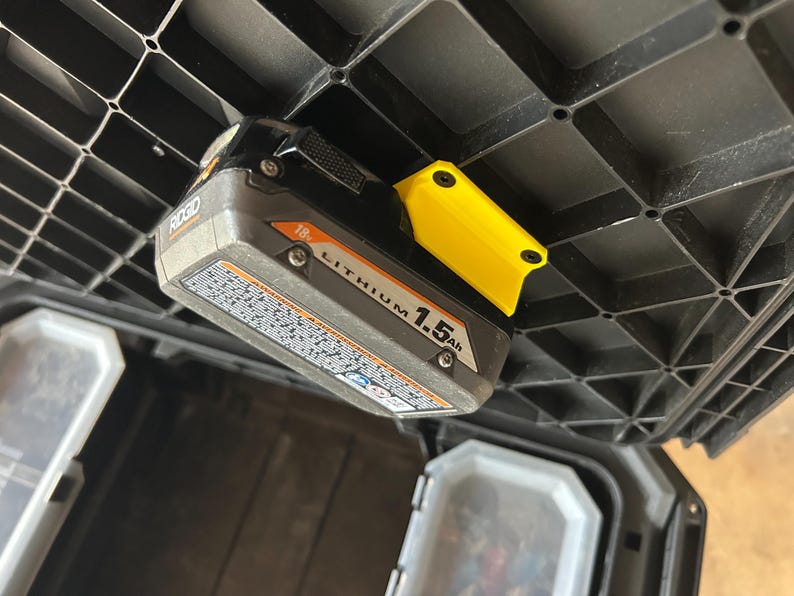 May include: A Ridgid 18V Lithium 1.5Ah battery pack with a yellow holder attached to a black plastic storage container. The battery is grey and black with white text.