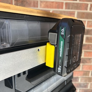 May include: A black and green Metabo 18V Li-ion 2.0Ah battery pack attached to a yellow and silver mounting bracket. The battery is mounted on a clear plastic storage container. The background is a brick wall.