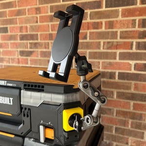 May include: Black tablet holder with an adjustable arm attached to a black and yellow storage container. The container has the words "TOUGHBUILT STACKTECH" printed on it. The background is a red brick wall.