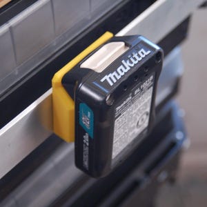 May include: A black Makita BL1021B 12V battery pack attached to a yellow holder. The battery has white text that reads "Makita" and a label with technical information. The holder is mounted on a metal surface.