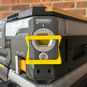 May include: A Toughbuilt 360° Pocket Level is attached to a gray and black toolbox. The level has a yellow frame and a circular dial with degree markings. The toolbox has the Toughbuilt logo and orange accents.