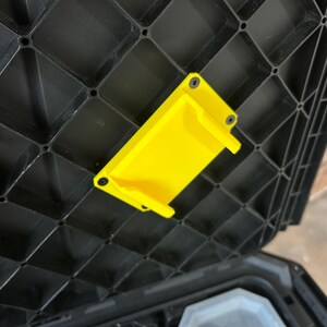 May include: A bright yellow plastic bracket is fixed to the inside of a black plastic storage container lid. The bracket is rectangular with two small tabs and is secured with black screws. The lid has a grid-like pattern.