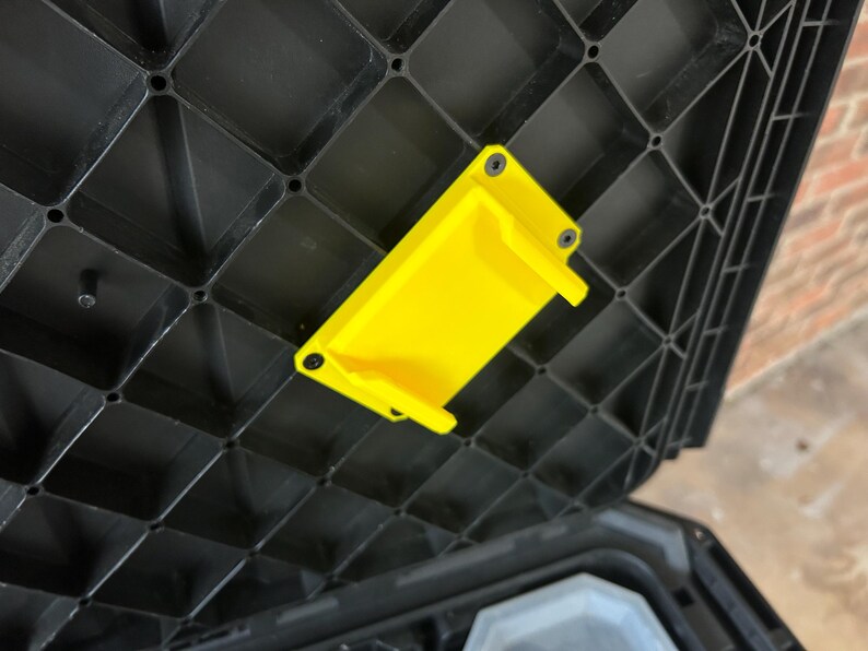 May include: A bright yellow plastic bracket is fixed to the inside of a black plastic storage container lid. The bracket is rectangular with two small tabs and is secured with black screws. The lid has a grid-like pattern.
