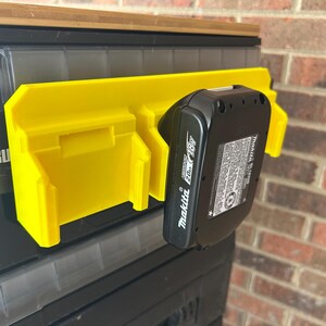 May include: A yellow plastic battery holder attached to a black storage container. A black Makita 18V battery is inserted into the holder. The holder is mounted on a black and gray storage container. The background is a brick wall.