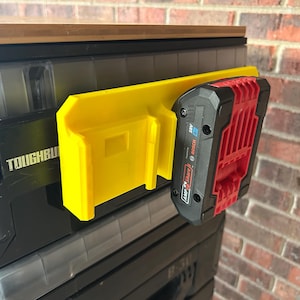 May include: A yellow 3D-printed battery holder attached to a black storage container. A red and black Bosch 18V battery is inserted into the holder. The container is labeled "TOUGHBUILT". The background is a brick wall.