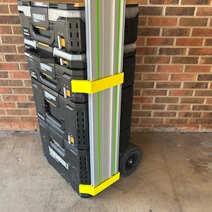 May include: A black rolling tool storage system with multiple compartments, stacked and secured. A silver and green guide rail is attached with yellow brackets. The system is on a concrete surface, with a brick wall in the background.