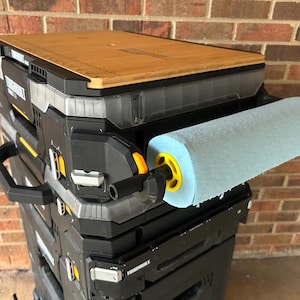 May include: A black and grey rolling tool chest with a wooden top and a blue paper towel roll attached. The tool chest has a handle and yellow accents. The background is a brick wall.