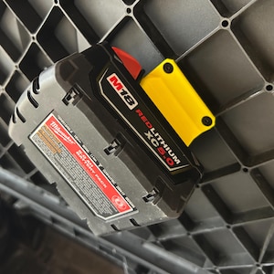 May include: A black and gray M18 Redlithium XC 5.0 battery pack with a yellow adapter, attached to a black grid-like surface. The battery pack has red accents and white text, including the Milwaukee brand name.