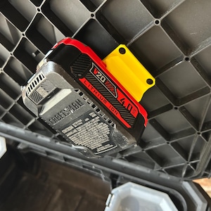 May include: A Craftsman V20 Lithium Ion 4.0 Ah battery pack, red and black, is mounted on a yellow holder. The battery is attached to a black plastic grid, likely part of a tool storage system.