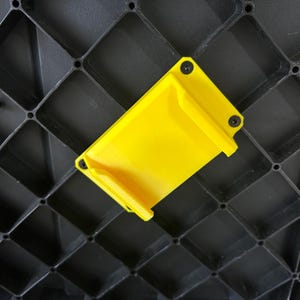 May include: A vibrant yellow plastic component, rectangular with rounded edges, secured to a black grid structure with small black screws. The component features a small lip on one side.