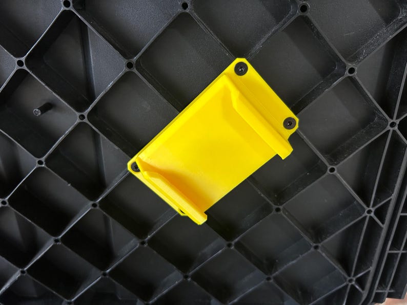 May include: A vibrant yellow plastic component, rectangular with rounded edges, secured to a black grid structure with small black screws. The component features a small lip on one side.