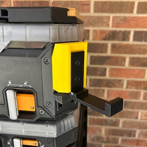 May include: A black and yellow tool organiser with a black hook extending from the side. The organiser has a clear top and is set against a brick wall.