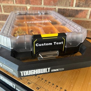 May include: A black and clear TOUGHBUILT STACKTECH storage container with a yellow accent and the words "Custom Text". The container has a clear lid and is filled with small, gold-colored objects. The container is sitting on a wooden surface.