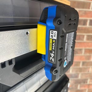 May include: A black and blue 24V MAX Lithium Ion 2.0 Ah battery pack attached to a yellow and blue holder. The battery is mounted on a metal shelf, with a brick wall in the background.