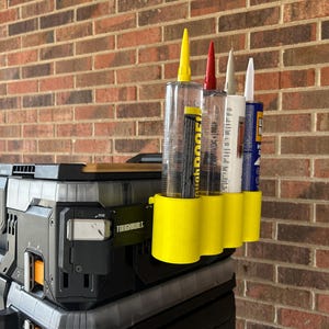 Peut inclure: Un support jaune pour tubes de calfeutrage contenant quatre tubes, chacun avec un embout de couleur différente. Le support est fixé à une boîte de rangement Toughbuilt noire et grise. En arrière-plan, un mur de briques rouges.