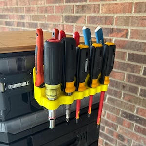 Peut inclure: Un support jaune pour outils contenant divers tournevis et un cutter. Les tournevis ont des manches noirs avec des accents jaunes et des tiges rouges. Le cutter est orange. Le support est monté sur un conteneur de stockage noir.