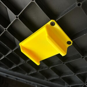 May include: A vibrant yellow plastic bracket, fixed with two black screws, is attached to a black, grid-patterned surface. The bracket's open-ended, rectangular shape suggests it's designed for holding or supporting an object.
