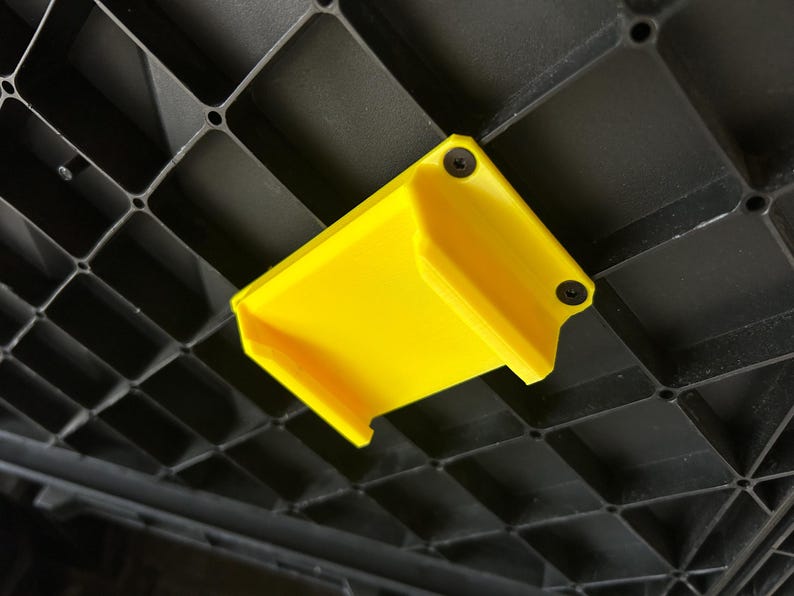 May include: A vibrant yellow plastic bracket, fixed with two black screws, is attached to a black, grid-patterned surface. The bracket's open-ended, rectangular shape suggests it's designed for holding or supporting an object.