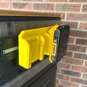 May include: A yellow battery holder with a black and yellow DeWalt 20V Lithium Ion battery attached. The holder is mounted on a black storage container. The word "TOUGHBUILT" is visible on the container.