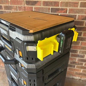 May include: A stack of black and gray toolboxes with a wooden top. A yellow battery holder and a black battery are attached to the side. The toolboxes are stacked on top of each other.