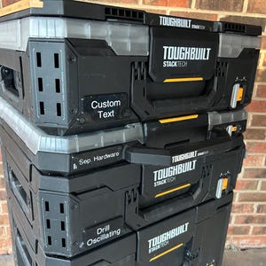 May include: A stack of black and grey ToughBuilt StackTech toolboxes. The boxes have clear plastic lids and yellow accents. Text on the boxes includes "Custom Text", "Sep. Hardware", and "Drill Oscillating". The ToughBuilt logo is also visible.