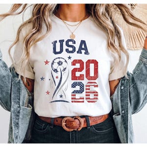 May include: A white t-shirt featuring a graphic design with the text USA 2026 in blue and red. The design includes a soccer ball, a stylized trophy shape, and small stars. The number 26 is filled with a red and white striped pattern.