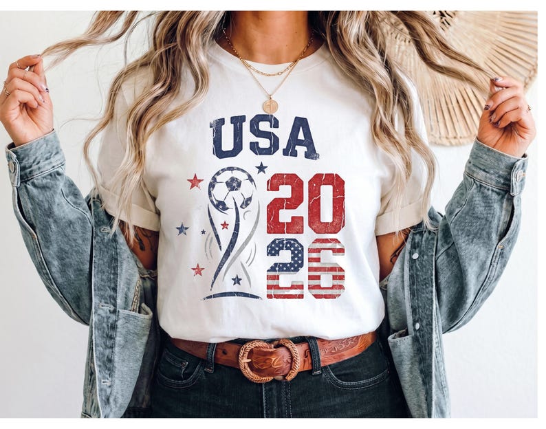 May include: A white t-shirt featuring a graphic design with the text USA 2026 in blue and red. The design includes a soccer ball, a stylized trophy shape, and small stars. The number 26 is filled with a red and white striped pattern.