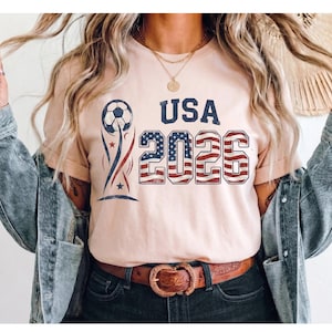 May include: A light peach-colored t-shirt featuring a graphic design with a soccer ball, the text USA, and the year 2026. The numbers 2026 are filled with a red, white, and blue American flag pattern, while the soccer ball has blue accents.