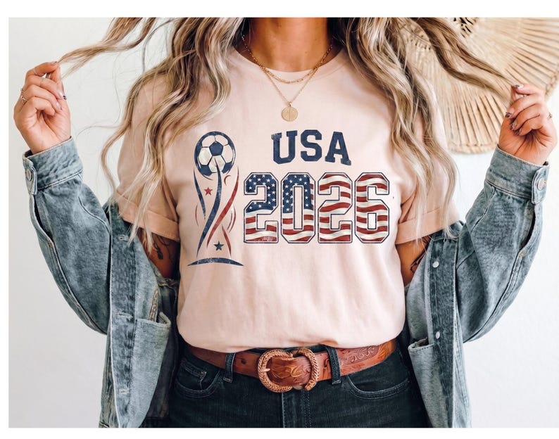 May include: A light peach-colored t-shirt featuring a graphic design with a soccer ball, the text USA, and the year 2026. The numbers 2026 are filled with a red, white, and blue American flag pattern, while the soccer ball has blue accents.