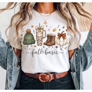May include: A white t-shirt featuring a graphic print of a green knit beanie, a leopard print tumbler with a bow, brown lace-up boots, and a white pumpkin with a brown bow, with the text fall basic written in black cursive script below.