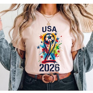 May include: A light peach t-shirt featuring a graphic print with the text USA 2026, a soccer ball, and a stylized trophy shape. The design includes colorful paint splatters in blue, yellow, and red, along with several small stars.