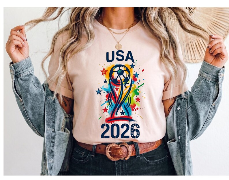 May include: A light peach t-shirt featuring a graphic print with the text USA 2026, a soccer ball, and a stylized trophy shape. The design includes colorful paint splatters in blue, yellow, and red, along with several small stars.