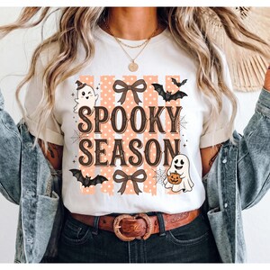 Puede incluir: Una camiseta blanca con el texto SPOOKY SEASON en letras marrones. El diseño incluye rayas verticales naranjas con lunares blancos, dos lazos marrones, dos fantasmas blancos, dos murciélagos negros y dos telarañas sobre fondo blanco.