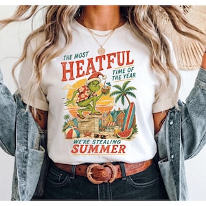May include: A white t-shirt featuring a graphic of a green creature wearing a Santa hat and a floral shirt, standing on a chimney. The text reads THE MOST HEATFUL TIME OF THE YEAR and WE'RE STEALING SUMMER, with beach elements like a surfboard.