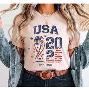 May include: A light peach-colored t-shirt featuring a distressed graphic with the text USA 2026 and EST. 2026. The design includes a soccer ball, stars, and stripes in red, white, and blue, layered over a stylized trophy shape with a worn texture.